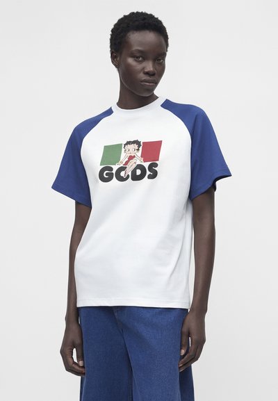 Άτομο που φοράει λευκό T-shirt με μπλε μανίκια, με τον κινούμενο χαρακτήρα Betty Boop πάνω από τα χρώματα της ιταλικής σημαίας και το κείμενο "GCDS", συνδυασμένο με μπλε παντελόνια.