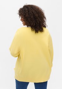 Donna con capelli ricci scuri che indossa un maglione giallo e jeans blu, in piedi di spalle contro uno sfondo bianco uniforme.