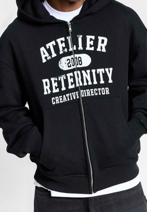 Person trägt einen schwarzen Kapuzenpullover mit Reißverschluss und weißem Text "ATELIER 2018 RETERNITY CREATIVE DIRECTOR" und hat die Hände in den Vordertaschen.