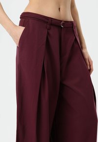 Burgundy wide-leg trousers featuring a high waistband with belt loops, front pleats, and side pockets. Smooth fabric texture designed for comfort.