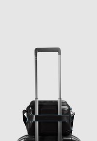 Zwarte leren tas vastgemaakt aan een koffer met wieltjes met een blauw accentbandje, voorzien van een ritssluiting aan de bovenzijde en een zijhandvat, op een grijze achtergrond.