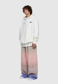 Överdimensionerad vit hoodie med ficka fram och broderad text; breda, pastellfärgade sweatpants i ombre som övergår från rosa till blått; vita sneakers.