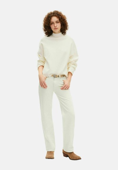 Giovane con capelli ricci indossa un maglione crema, pantaloni chiari con una cintura e stivali alla caviglia in suede marrone, in piedi con le mani nelle tasche.