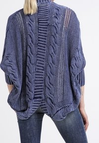 Poncho de punto en azul oscuro con un patrón de trenzas, bordes acanalados y un diseño abierto y drapeado. Textura suave y forma relajada.