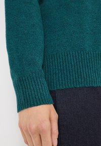 Maglione in maglia teal con polsini e orlo a costine, che mette in risalto una texture morbida e una vestibilità aderente. Il design sottolinea calore e comfort.