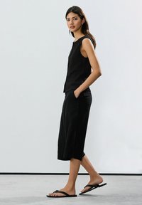 Black sleeveless textured top paired with loose black culottes and flat sandals. Model stands with one hand in pocket against a plain backdrop.