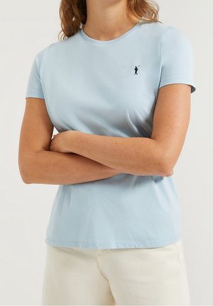 Kvinde iført en lyseblå kortærmet skjorte med en lille mørk golfspillerlogo på venstre bryst, med krydsede arme, stående foran en ensfarvet baggrund.