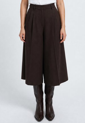 Woman wearing high-waisted, wide-leg dark brown culottes paired with knee-high dark brown leather boots on a light background.