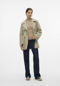 Trench coat beige de doble botonadura con grandes solapas, mangas largas y epaulettes en los hombros, combinado con jeans de pierna ancha de color azul oscuro y zapatillas deportivas.