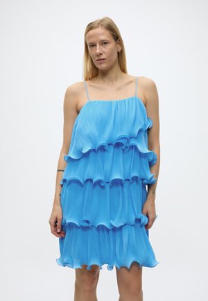 Frau trägt ein ärmelloses, mehrlagiges blaues Kleid mit dünnen Trägern und steht vor einem schlichten weißen Hintergrund.