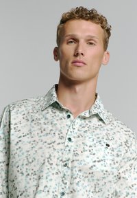 Lichtblauw en witte button-up blouse met een geometrisch patroon, gestructureerde kraag en teal knopen. De stof lijkt glad en lichtgewicht.
