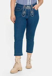 Jeans i denim med broderade hängslen, mörkblå färg, kort längd och framfickor, kombinerade med beiga snörstövlar.