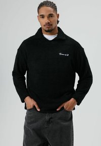 Pull-over en polaire noire avec une surface texturée et un col montant. Comprend un logo blanc brodé sur la poitrine et une coupe décontractée.