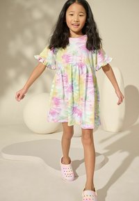 Šarena tie-dye haljina u pastelnim nijansama ružičaste, zelene i žute, s naboranim rukavima i opuštenim krojem, uparena s laganim crocsicama.