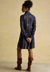 Merineelik kuldse ja punase graafilise mustriga, pikkade varrukatega, kaelusega, kehakumerustega ja laieneva seeliku lõikega. Kombineeritud pruuni luksusliku nahast põlvini ulatuvate saapadega.