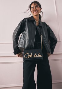Juoda odinė striukė su apykakle, derinama su juoda rankine, ant kurios auksiniu užrašu „Ooh lala“ ir grandinėlės diržu, dėvima ant juodų kelnių.