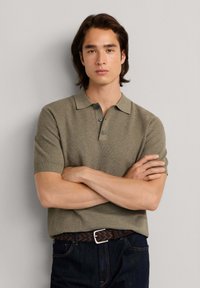 Jeune homme aux cheveux mi-longs portant un polo à manches courtes vert olive et un jean foncé, debout les bras croisés.