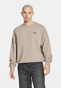 Beżowy długi rękaw bluza z okrągłym dekoltem i prążkowanymi mankietami. Posiada czarny nadruk logo na lewej piersi. Wykonana z miękkiej bawełny.