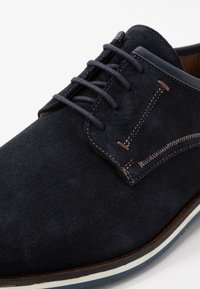 Les chaussures en daim bleu marine présentent des lacets, un design cousu avec un accent contrastant, et une semelle à rayures blanches pour une finition propre et moderne.