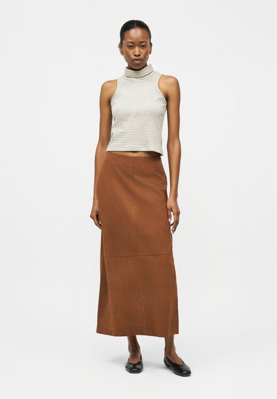 Γυναίκα που φοράει ριγέ τοπ χωρίς μανίκια και με υψηλό λαιμό, καφέ μακριά φούστα και μαύρα επίπεδα παπούτσια, στέκεται σε απλό ανοιχτό φόντο.