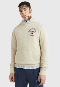 Felpa beige con cappuccio, caratterizzata da tasca frontale, logo ricamato e polsini a coste. Indossata sopra una camicia azzurra con bottoni.
