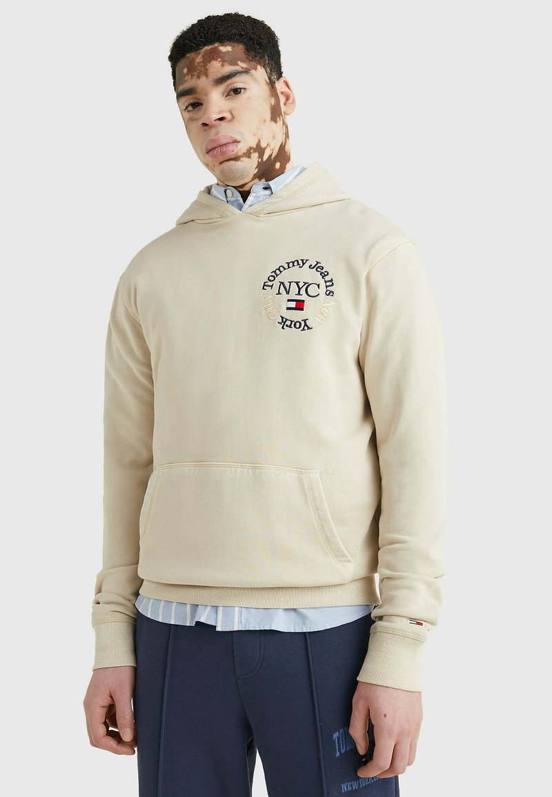 Felpa beige con cappuccio, caratterizzata da tasca frontale, logo ricamato e polsini a coste. Indossata sopra una camicia azzurra con bottoni.