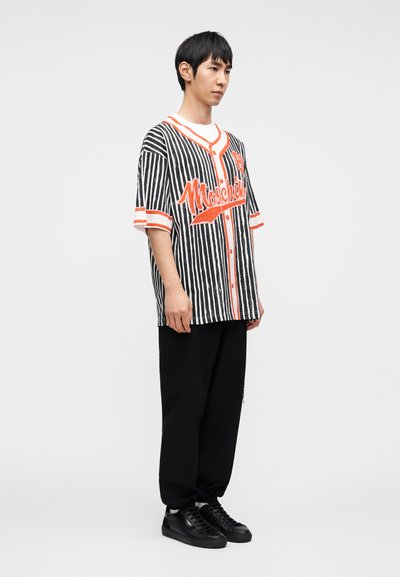 Ριγέ μπλουζάκι μπέιζμπολ με κοντά μανίκια σε μαύρο και άσπρο, με κόκκινες λεπτομέρειες και γράμματα "Moschino", σε συνδυασμό με χαλαρά μαύρα παντελόνια και αθλητικά παπούτσια.