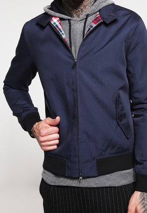 Marineblauwe bomberjack met een rits aan de voorkant, zwarte geribbelde manchet en een rood geruit voering. Draagt over een grijze hoodie.