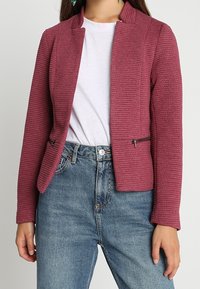 Texturerad bordeauxjacka med krage, utrustad med två sidofickor med dragkedja, kombinerad med en vit t-shirt och blå denimjeans.