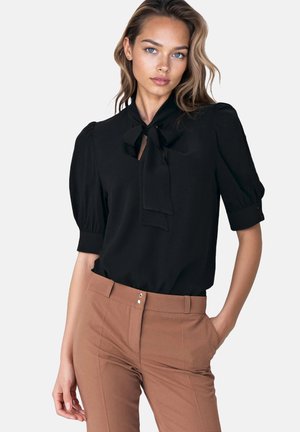 Kobieta w czarnej bluzce z kołnierzykiem w formie kokardy i bufiastymi rękawami, włożonej w brązowe, eleganckie spodnie o wysokim stanie, z ręką w kieszeni.