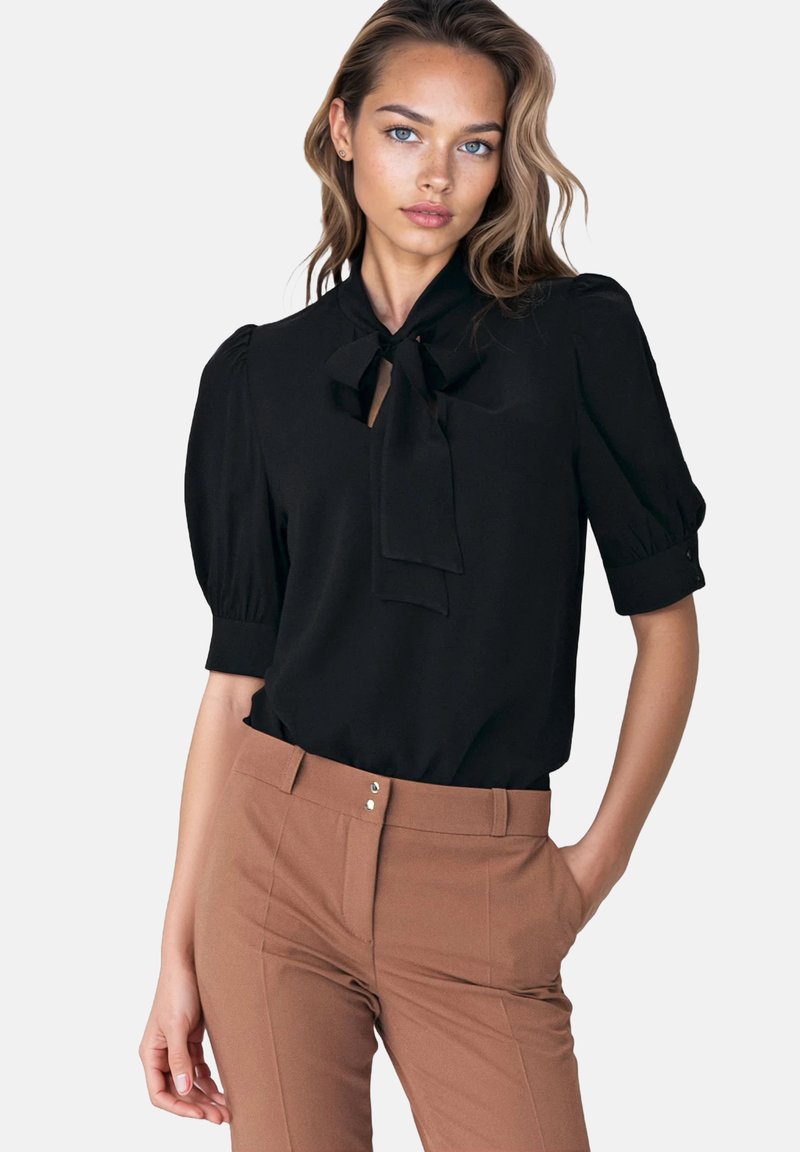 Kobieta w czarnej bluzce z kołnierzykiem w formie kokardy i bufiastymi rękawami, włożonej w brązowe, eleganckie spodnie o wysokim stanie, z ręką w kieszeni.