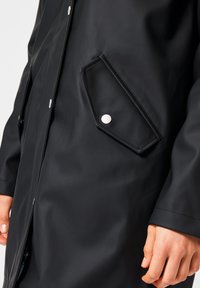 Imperméable noir avec une finition brillante, poches à pression et coupe droite avec des détails de couture visibles sur les poignets.