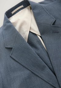 Blazer a righe blu scuro, con rever a punta, tessuto testurizzato e fodera interna color crema. Cuciture visibili e dettagli delle tasche.