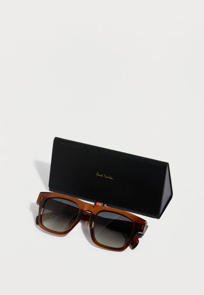 Gafas de sol rectangulares marrones con lentes degradados, colocadas junto a un estuche triangular negro con texto dorado en relieve.