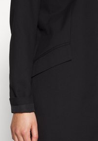 Blazer negro con cuello de solapa, tejido texturizado y detalle de puño con botones. El diseño presenta un corte elegante y entallado.