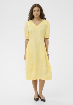 Nő áll, térdig érő sárga ruhát visel, melynek puffos rövid ujjai vannak, és fekete hegyes orrú magas sarkú cipőt, fehér háttér előtt.