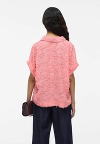 Roze gekante blouse met een kraag en korte mouwen, met een gelaagd, rafelig ontwerp. Gecombineerd met donkere, losse broek.