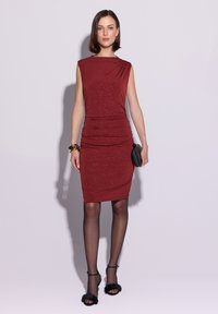 Czerwona marszczona sukienka wykonana z miękkiej tkaniny, z wysokim kołnierzem, drapowaniem na ramionach i subtelnym błyskiem. Noszona z przezroczystymi czarnymi rajstopami i sandałami na paskach.
