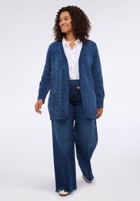 Cárdigan de manga larga en punto texturizado de color azul oscuro, con frente abierto, combinado con una camisa blanca y pantalones de denim de pierna ancha, creando una silueta relajada.