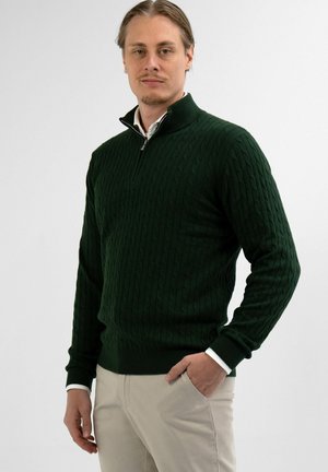 Donkergroene kabelgebreide sweater met een ritskraag, geribbelde manchetten en zoom. In combinatie met lichtbeige broek, staande tegen een neutrale achtergrond.