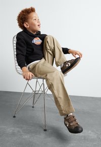 Bruna sneakers med vita stjärnaccenter, beige byxor och en svart hoodie med en färgglad logotyp, sittande på en trådstol.