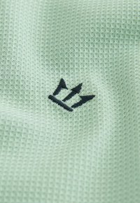 Tissu vert clair texturé avec un petit logo brodé noir en forme de trident au centre.