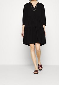 Schwarzes, gestuftes Kleid mit V-Ausschnitt und Dreiviertelärmeln. Kombiniert mit burgunderfarbenen, gekreuzten Sandalen. Schlichte Textur, knielanges Design.