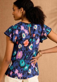 Zila rakstainā blūze ar īsām volānu piedurknēm, kas rotāta ar krāsainiem punktiem rozā, zaļā un orandžā krāsā, un ar mazu atslēgas caurumu aizmugurē.