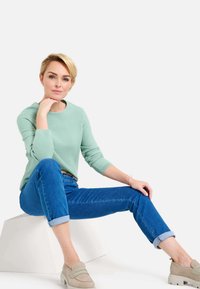 Maglione verde menta lavorato a maglia, jeans blu con risvolto e scarpe beige senza lacci. Modella seduta su un blocco geometrico bianco, sfondo neutro.
