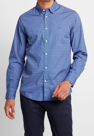 Homme portant une chemise bleue ajustée à motifs avec des manches longues et un pantalon foncé, debout devant un fond uni.