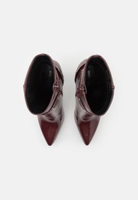 Bottines en cuir verni bourgogne avec un bout pointu et une fermeture éclair sur le côté, ornées d'accents métalliques et d'une texture lisse et brillante.