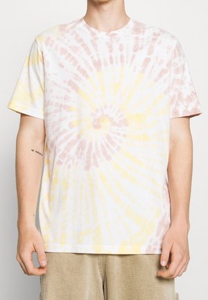 Osoba nosząca krótką koszulkę z krótkim rękawem z wzorem tie-dye z spiralnymi motywami w jasnobrązowym i bladżółtym kolorze, do tego beżowe spodnie sztruksowe.