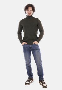 Maglione a collo alto verde scuro con texture a coste, abbinato a jeans blu e sneakers in pelle marrone con accenti chiari e lacci.