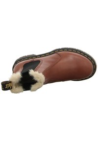 Bottine en cuir marron avec panneaux latéraux élastiques noirs, doublure en shearling, surpiqûres jaunes sur la semelle noire, et languette de traction brandée au talon.